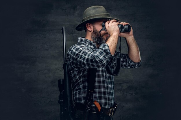 Retrato de estudio de un cazador barbudo que usa una camisa de lana a cuadros mirando a través de binoculares, sostiene un rifle.