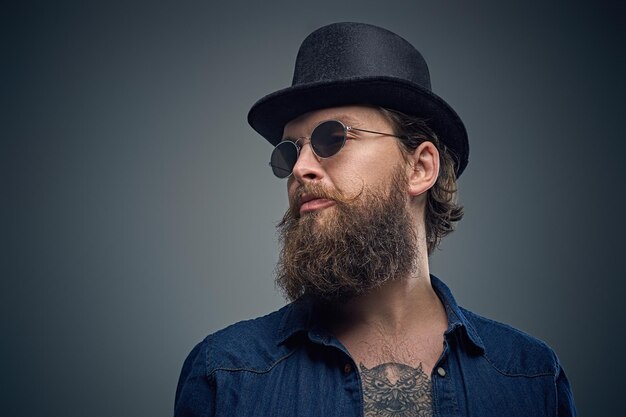 Retrato de estudio de un elegante hombre barbudo con un tatuaje en el pecho, vestido con una camisa de mezclilla, un sombrero cilíndrico y gafas de sol aisladas en un fondo gris.