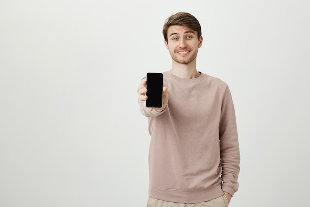 Retrato de estudio de feliz atractivo modelo masculino europeo mostrando smartphone mientras sonríe y sostiene una mano en el bolsillo