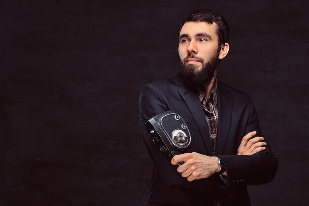 Retrato de estudio de un fotógrafo barbudo con un traje clásico que sostiene una cámara retro, de pie con los brazos cruzados.