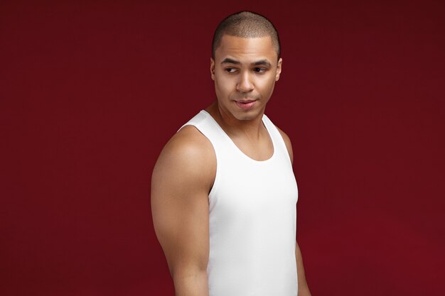 Retrato de estudio de fresco joven afroamericano de 20 años de edad con camiseta blanca sin mangas posando aislado. Chico guapo serio de piel oscura con hombros musculosos descansando en el interior