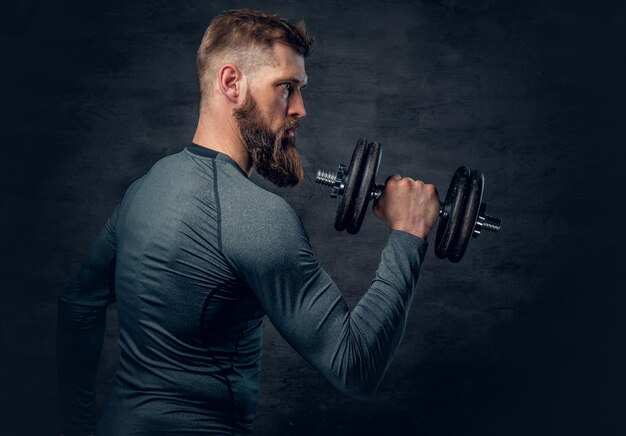 Retrato de estudio de un hombre barbudo deportivo vestido con ropa deportiva gris que sostiene pesas.