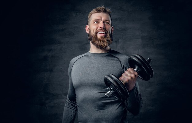 Retrato de estudio de un hombre barbudo deportivo vestido con ropa deportiva gris que sostiene pesas.