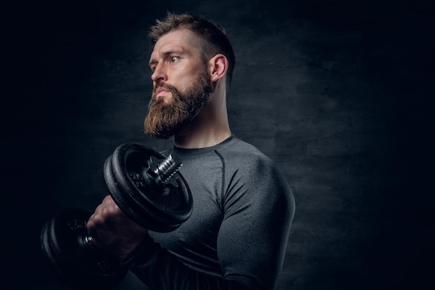 Retrato de estudio de un hombre barbudo deportivo vestido con ropa deportiva gris que sostiene pesas.