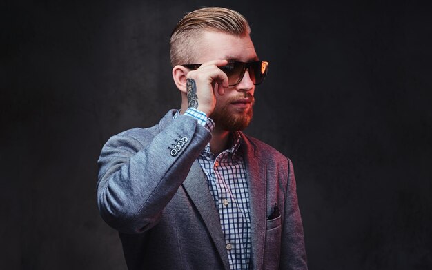 Retrato de estudio de hombre barbudo pelirrojo vestido con traje y gafas de sol sobre fondo gris.