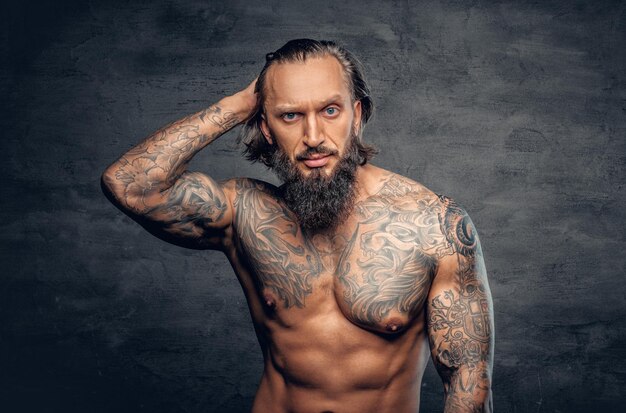 Retrato de estudio de un hombre barbudo tatuado y sin camisa sobre un fondo de viñeta gris oscuro.