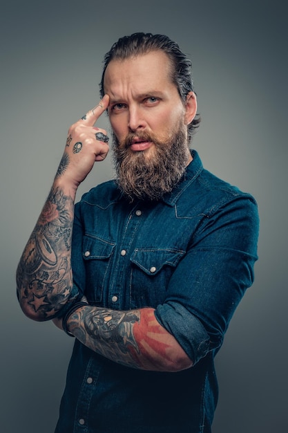 Retrato de estudio de un hombre barbudo con tatuajes en los brazos, vestido con una camisa de mezclilla aislada en un fondo de viñeta gris.