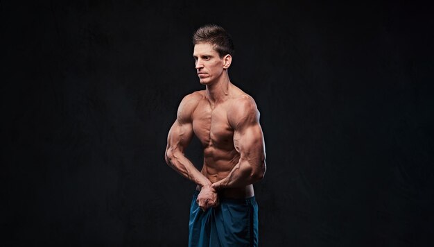 El retrato de estudio de un hombre sin camisa con músculo ectomorfo muestra sus bíceps sobre un fondo gris oscuro.