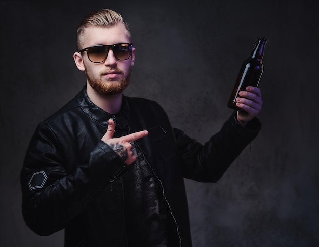 Retrato de estudio de un hombre elegante con barba rubia sostiene una botella con cerveza artesanal.