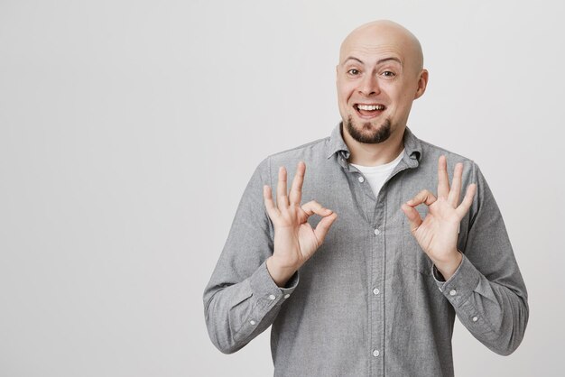 Retrato de estudio de un hombre europeo alegre, calvo y apuesto con barba que muestra un gesto bueno o genial mientras sonríe ampliamente aprobando o estando de acuerdo con algo sobre un fondo gris