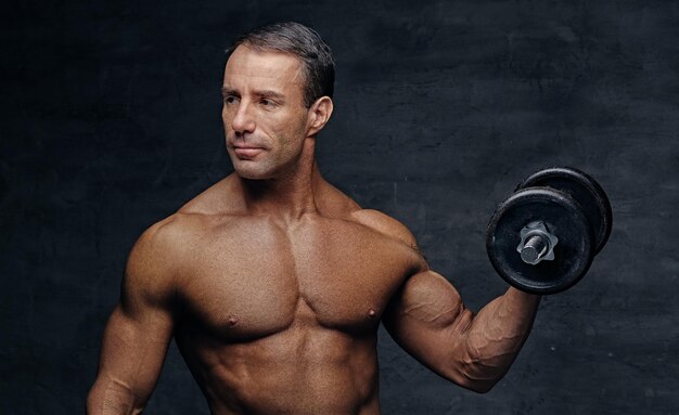 Retrato de estudio de un hombre de mediana edad musculoso sin camisa sostiene un dumbbel aislado en un fondo de viñeta gris.