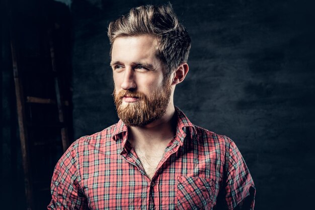 Retrato de estudio de un hombre rubio con barba vestido con una camisa de lana roja sobre un fondo gris oscuro.