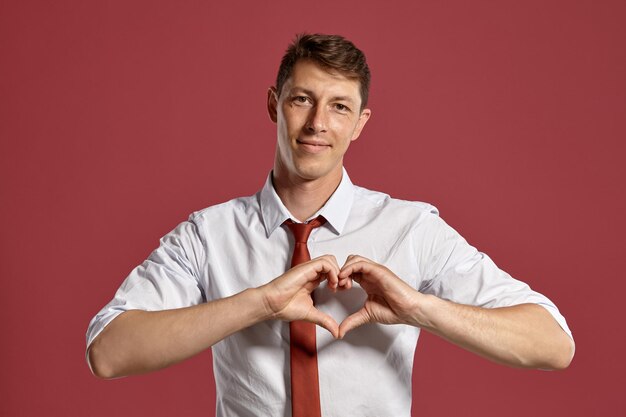 Retrato de estudio de un joven atlético con un clásico pantalones blanco y corbata roja, mostrando un corazón doblado de sus dedos mientras posa sobre un fondo rosa. Corte de pelo con estilo. Concepto de emociones sinceras. Co