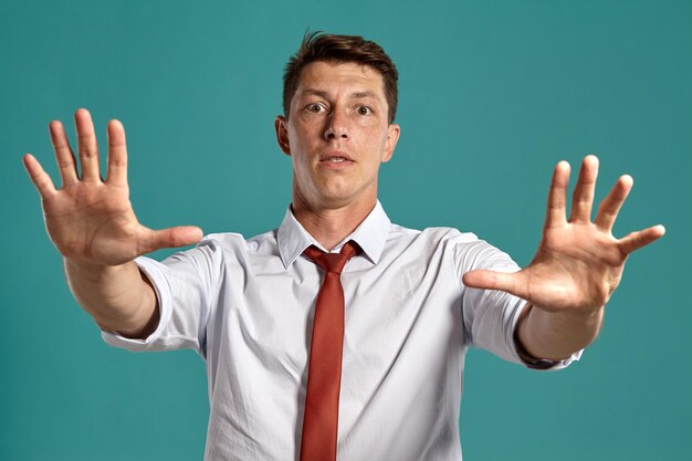 Retrato de estudio de un joven majestuoso con una camisa blanca clásica y una corbata roja que actúa como detener a alguien y mirar a la cámara mientras posa sobre un fondo azul. Corte de pelo con estilo. emoción sincera