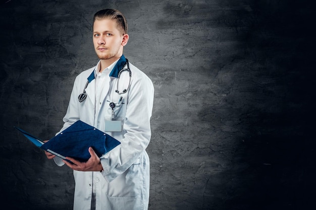 El retrato de estudio de un joven médico sostiene un portapapeles azul sobre fondo gris.