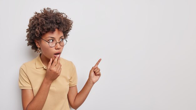 El retrato de estudio de una mujer de cabello rizado asombrada mantiene la mandíbula caída por el asombro, mira fijamente los ojos saltones, usa anteojos redondos, una camiseta beige informal aislada sobre fondo blanco. Dios mío, mira esto.