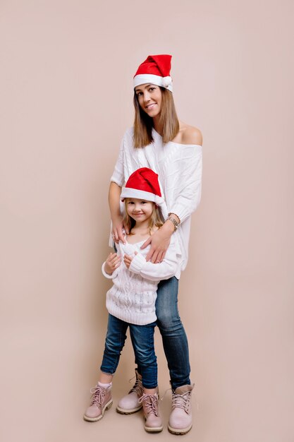 Retrato de estudio de mujer joven y atractiva con su pequeña hija vistiendo suéteres blancos y sombreros de Navidad