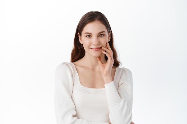 Retrato de estudio de una mujer joven que muestra una emoción tranquila y relajada sonriendo suavemente mirando a la cámara tocando la cara natural con maquillaje desnudo de pie contra el fondo blanco