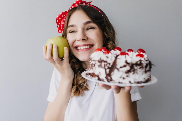 Retrato de estudio de mujer rizada alegre posando con pastel y manzana Niña sonriente con cinta roja en el cabello sosteniendo sabrosa manzana verde y pastel de cumpleaños