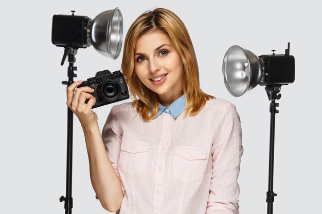 Retrato de estudio de mujer rubia positiva con cámara fotográfica con equipo fotográfico en el fondo.