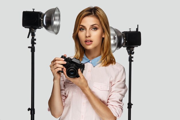 Retrato de estudio de mujer rubia positiva con cámara fotográfica con equipo fotográfico en el fondo.