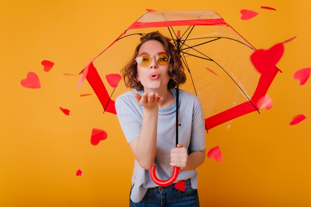 Retrato de estudio de niña blanca con gafas de sol disfrutando del día de San Valentín. Foto interior de una mujer increíble posando bajo el paraguas con expresión de la cara de besos.