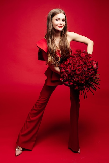 Retrato de estudio de vista lateral de una hermosa joven morena con el pelo largo y un vestido rojo de moda Ella está sonriendo a un gran ramo de rosas rojas en sus manos Color rojo total Aislada sobre fondo rojo
