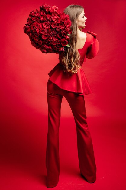 Retrato de estudio de vista lateral de una hermosa joven morena con el pelo largo y un vestido rojo de moda Ella está sonriendo a un gran ramo de rosas rojas en sus manos Color rojo total Aislada sobre fondo rojo