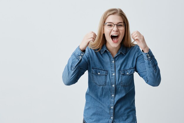 Retrato de la excitada modelo femenina con cabello rubio, gafas, camisa de mezclilla, aprieta los puños con placer, grita de felicidad, celebra la victoria, tiene un gran triunfo. Concepto de éxito