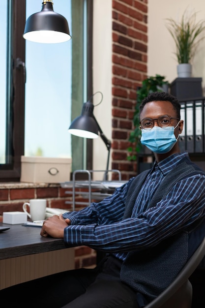 Retrato de un exitoso empleado de inicio con máscara facial covid sentado en el escritorio mirando la cámara. Empresario afroamericano con anteojos posando confiado sentado en la oficina de negocios.