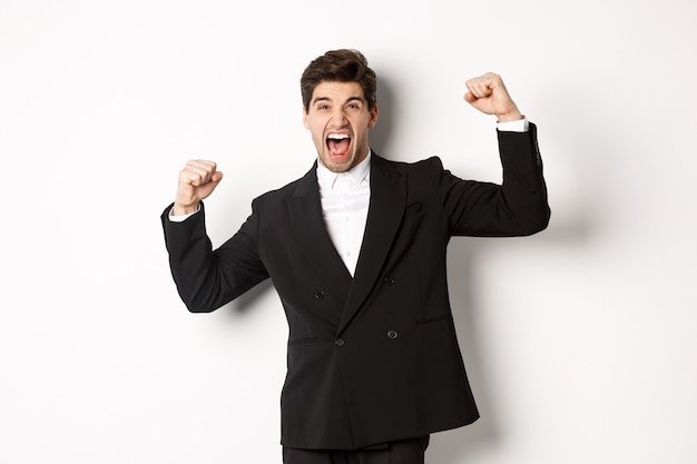 Foto gratuita retrato de exitoso hombre de negocios en traje negro, convertirse en campeón, levantando las manos y gritando que sí, triunfando y celebrando la victoria, de pie contra el fondo blanco.