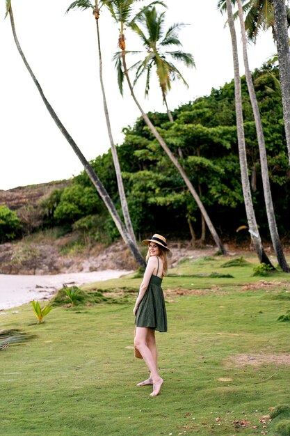 Retrato exótico de verano de una mujer rubia bonita posando en una isla exótica, con vestido y sombrero de paja. Vacaciones de lujo en la isla.