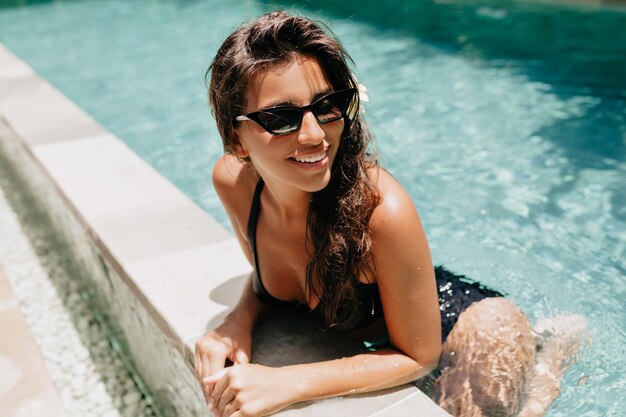 Retrato exterior de adorable linda dama con cabello oscuro divirtiéndose en la piscina en un día cálido y soleado