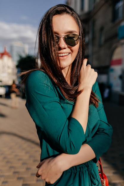 Retrato exterior de una encantadora mujer sonriente con gafas de sol y vestido verde posando en la cámara en una calle soleada de verano