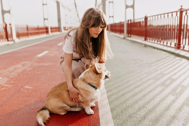 El retrato exterior de una joven dama bonita con el pelo largo, vestida con una camiseta blanca y una falda, camina por el puente con su encantador perro