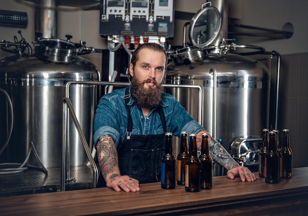 Retrato de un fabricante masculino tatuado y barbudo que presenta cerveza en la microcervecería.