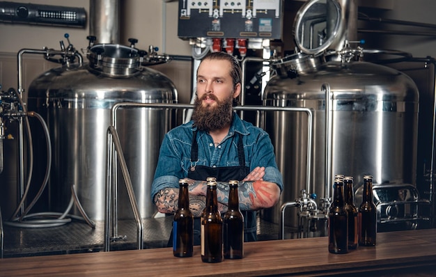 Foto gratuita retrato de un fabricante masculino tatuado y barbudo que presenta cerveza en la microcervecería.