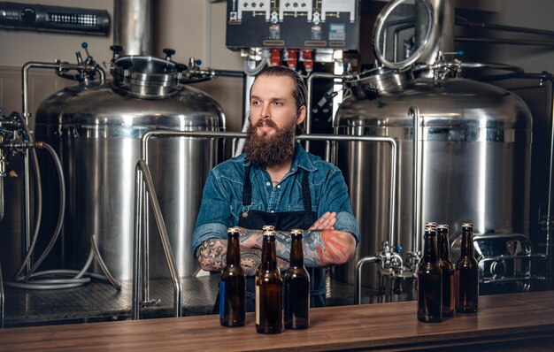 Retrato de un fabricante masculino tatuado y barbudo que presenta cerveza en la microcervecería.