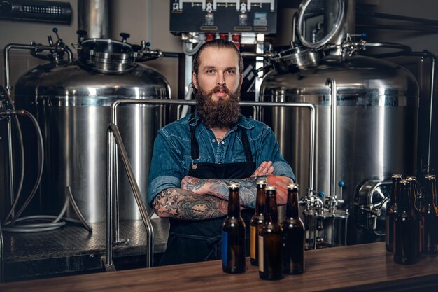 Retrato de un fabricante masculino tatuado y barbudo que presenta cerveza en la microcervecería.