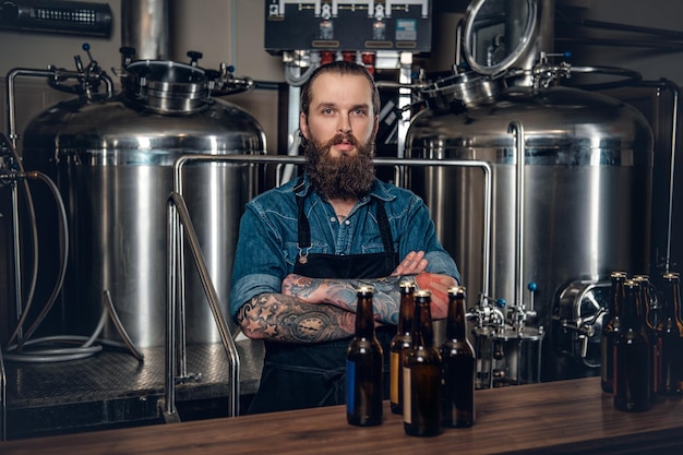 Retrato de un fabricante masculino tatuado y barbudo que presenta cerveza en la microcervecería.