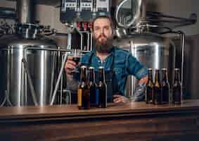 Foto gratuita retrato de un fabricante masculino tatuado y barbudo que presenta cerveza en la microcervecería.