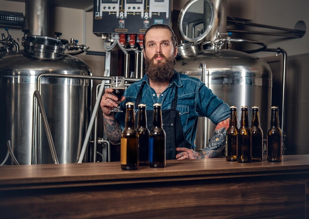 Retrato de un fabricante masculino tatuado y barbudo que presenta cerveza en la microcervecería.