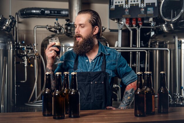 Retrato de un fabricante masculino tatuado y barbudo que prueba cerveza en la microcervecería.