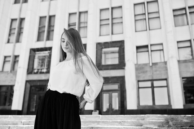 Retrato de una fabulosa joven exitosa con blusa blanca y amplios pantalones negros posando en las escaleras con un enorme edificio blanco al fondo Foto en blanco y negro
