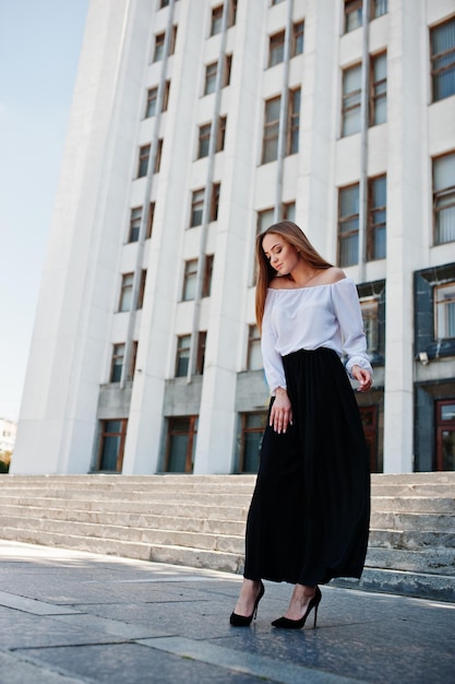 Retrato de una fabulosa joven exitosa con blusa blanca y amplios pantalones negros posando en las escaleras con un enorme edificio blanco al fondo