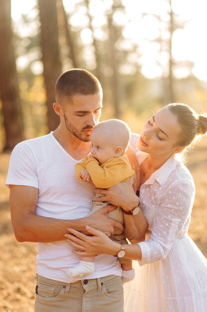 Retrato de familia atractiva joven con hijo pequeño, posando en el hermoso bosque de pinos de otoño en un día soleado. Hombre guapo y su bella esposa morena