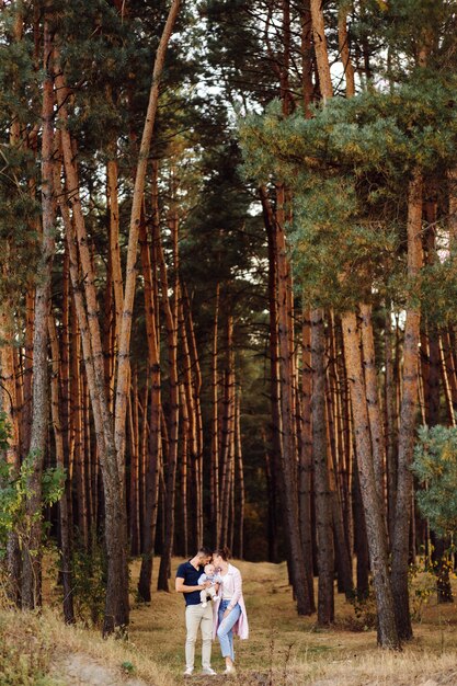 Retrato de familia atractiva joven con hijo pequeño, posando en el hermoso bosque de pinos de otoño en un día soleado. Hombre guapo y su bella esposa morena