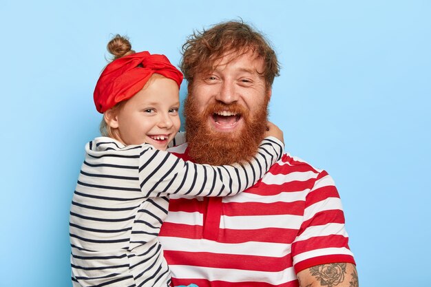 Retrato de familia de hija feliz viste diadema roja y jersey de rayas, abraza a padre encantado con barba espesa de jengibre y cabello rizado, se aman mucho