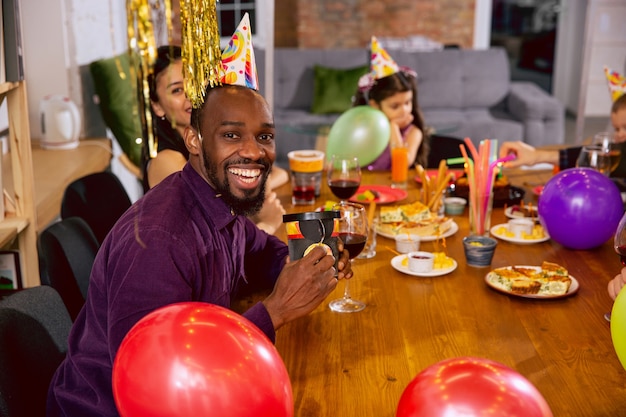 Retrato de familia multiétnica feliz celebrando un cumpleaños en casa. Gran familia comiendo bocadillos y bebiendo vino mientras saluda y se divierte a los niños. Celebración, familia, fiesta, concepto de hogar.