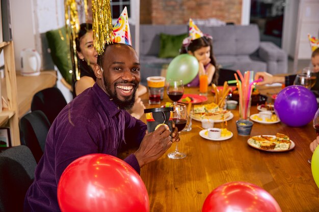 Retrato de familia multiétnica feliz celebrando un cumpleaños en casa. Gran familia comiendo bocadillos y bebiendo vino mientras saluda y se divierte a los niños. Celebración, familia, fiesta, concepto de hogar.
